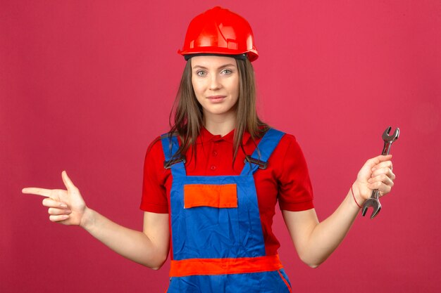 Mujer joven en uniforme de construcción y casco de seguridad rojo sonriendo y señalando con el dedo hacia un lado y sosteniendo una llave ajustable sobre fondo rosa oscuro