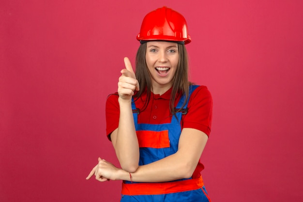 Mujer joven en uniforme de construcción y casco de seguridad rojo sonriente y feliz mirando apuntando a la cámara de pie sobre fondo rosa oscuro