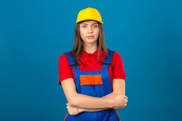 Mujer joven en uniforme de construcción y gorra amarilla de pie con los brazos cruzados mirando a la cámara con cara seria sobre fondo azul.