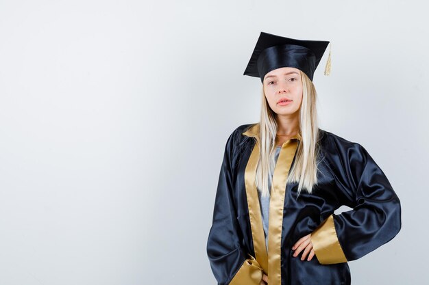 Mujer joven en uniforme de posgrado posando con la mano en la cintura y mirando encantador