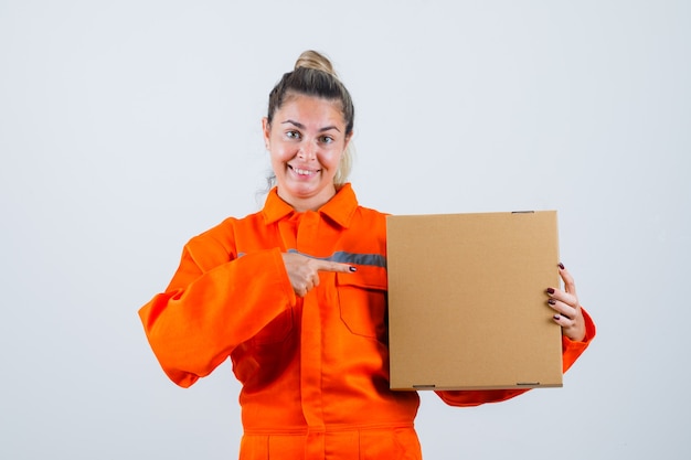 Mujer joven en uniforme de trabajador apuntando a la caja y mirando contento, vista frontal.