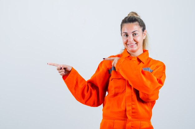 Mujer joven en uniforme de trabajador apuntando a un lado mientras sonríe y mira alegre, vista frontal.