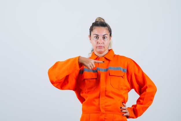 Mujer joven en uniforme de trabajador apuntando a un lado y mirando serio, vista frontal.