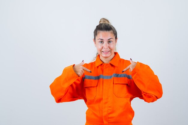 Mujer joven en uniforme de trabajador apuntando a sí misma y mirando dudosa, vista frontal.