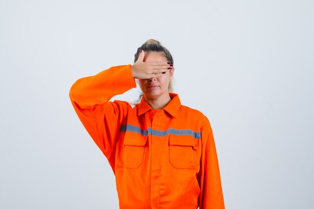 Mujer joven en uniforme de trabajador cubriendo los ojos con la mano y mirando emocionado, vista frontal.