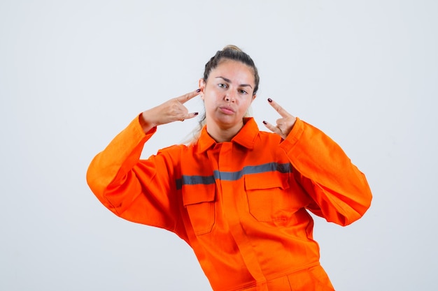 Mujer joven en uniforme de trabajador haciendo rock in roll símbolo con las manos en alto y mirando seguro, vista frontal.