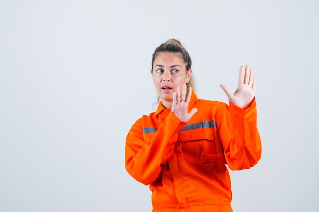 Foto gratuita mujer joven en uniforme de trabajador levantando las manos para defender y mirando asustado, vista frontal.