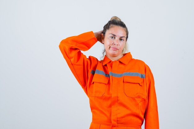 Mujer joven en uniforme de trabajador sosteniendo la mano detrás de la cabeza y mirando pensativo, vista frontal.