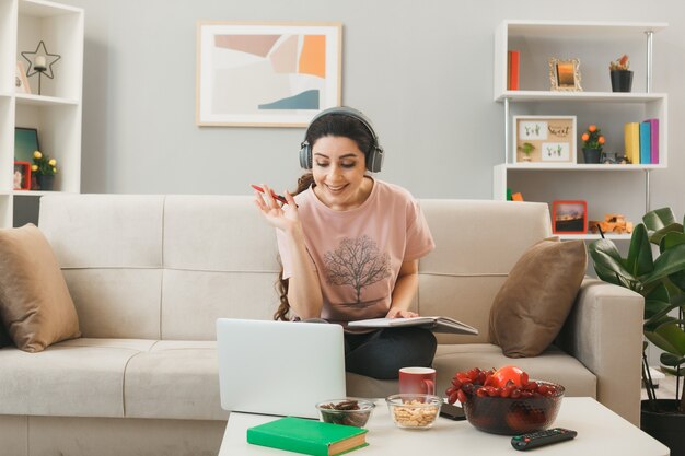 Mujer joven usando audífonos sosteniendo la pluma con el libro portátil usado sentado en el sofá detrás de la mesa de café en la sala de estar