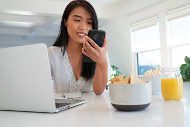 Mujer joven usando una computadora portátil y un teléfono inteligente mientras come chips de tortilla