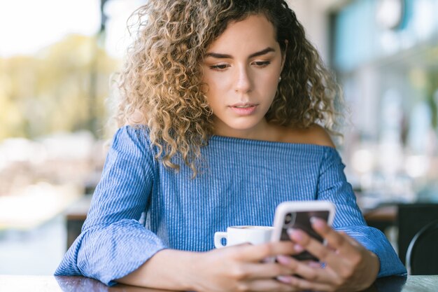 Mujer joven usando un teléfono móvil y bebiendo una taza de café en una cafetería.