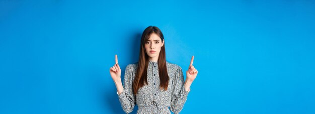 Mujer joven vacilante y preocupada frunciendo el ceño señalando con el dedo hacia arriba con cara triste de pie en backgro azul