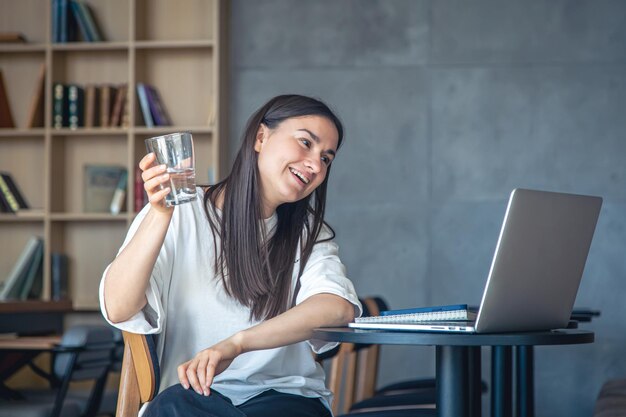 Mujer joven con un vaso de agua frente a una computadora portátil