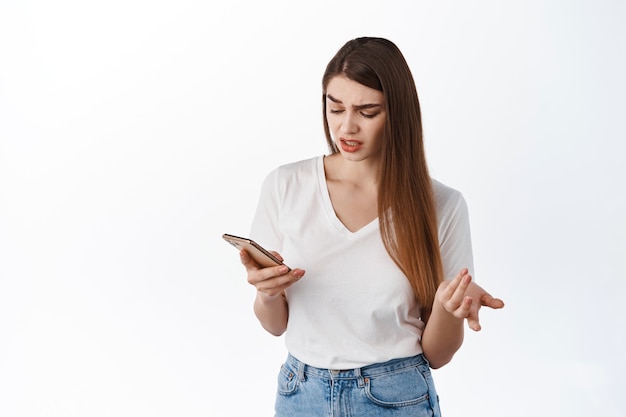 Mujer joven se ve confundida en la pantalla del móvil, leyendo un mensaje extraño, no entiendo algo en el teléfono, mira desconcertado al teléfono inteligente, de pie sobre una pared blanca