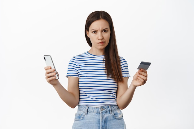 Foto gratuita la mujer joven se ve dudosa, frunce las cejas y mira vacilante, sospechosa a la cámara, sostiene un teléfono inteligente con tarjeta de crédito, concepto de compras en línea y dinero.