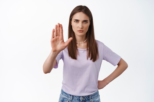 La mujer joven se ve seria muestra que no, levantando la palma para bloquear o prohibir algo, prohibir la entrada al lugar, de pie en blanco