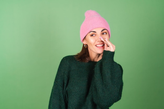 Mujer joven en verde en un cálido suéter acogedor y un sombrero rosa dice un secreto, susurros, chismes