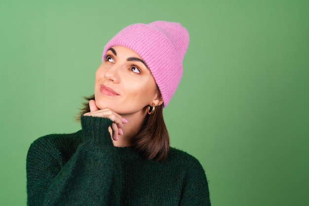 Mujer joven en verde en un cálido suéter acogedor y un sombrero rosa sonríe tiernamente, mira pensativamente a un lado, inspirado