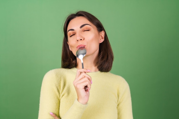 Mujer joven en verde con un suéter amarillo con apetito come de una cucharada, disfrutando