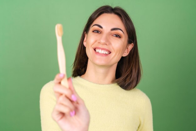 Mujer joven en verde con un suéter amarillo sostiene un cepillo de dientes de madera ecológico, cuidando la naturaleza y un diente