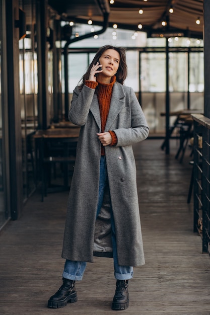 Foto gratuita mujer joven vestida con abrigo gris y caminando por la calle y usando el teléfono