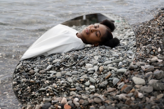 Mujer joven vestida de blanco posando con espejo en rocas junto al agua