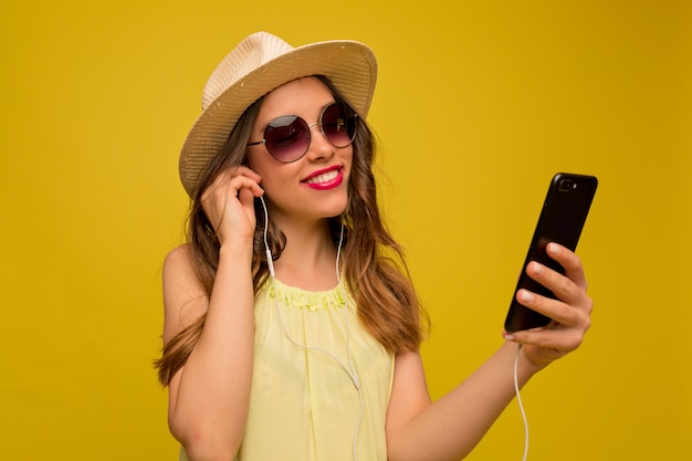 Mujer joven en vestido amarillo con sombrero y gafas de sol escuchando música