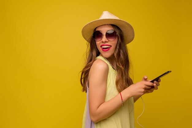 Mujer joven en vestido amarillo con sombrero y gafas de sol con smartphone