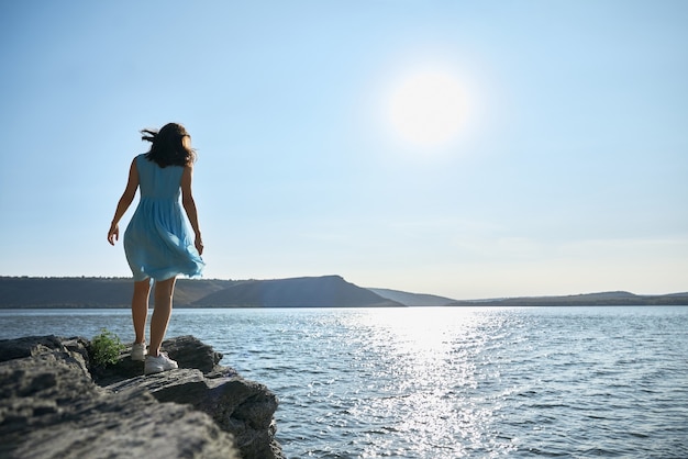 Mujer joven en vestido azul caminando por la orilla del río