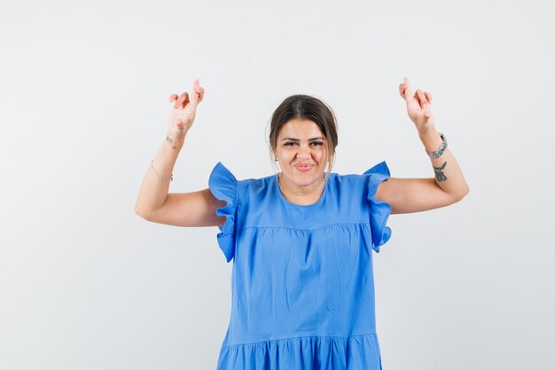 Mujer joven en vestido azul manteniendo los dedos cruzados y mirando feliz