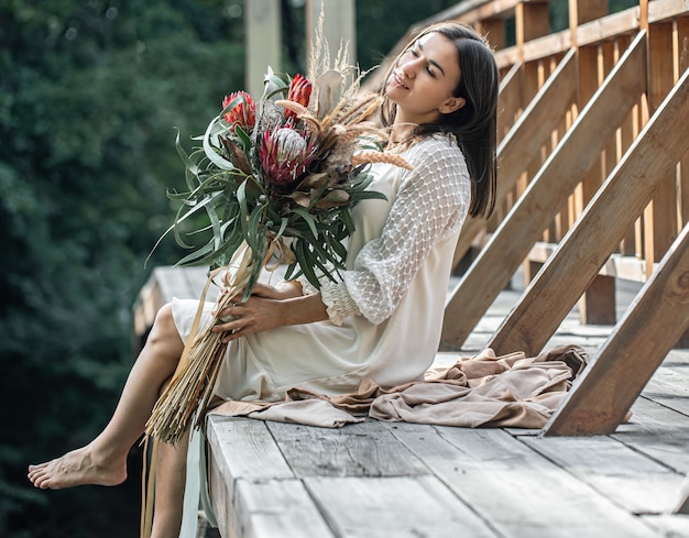 Una mujer joven con un vestido blanco se sienta en un puente de madera con un ramo de flores exóticas.