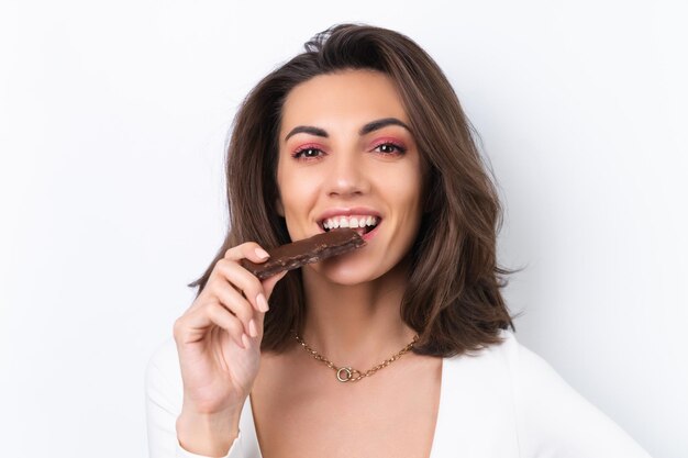 Mujer joven en un vestido de cóctel cadena de oro maquillaje rosa primavera brillante sobre un fondo blanco sostiene una deliciosa barra de chocolate cereal saludable