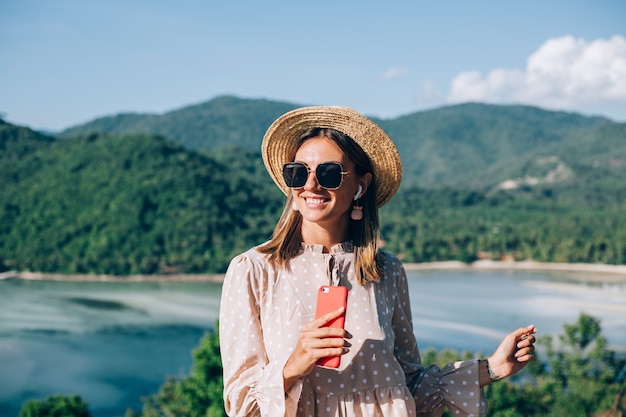 Mujer joven en vestido lindo de verano, sombrero de paja y gafas de sol bailando con smartphone en mano