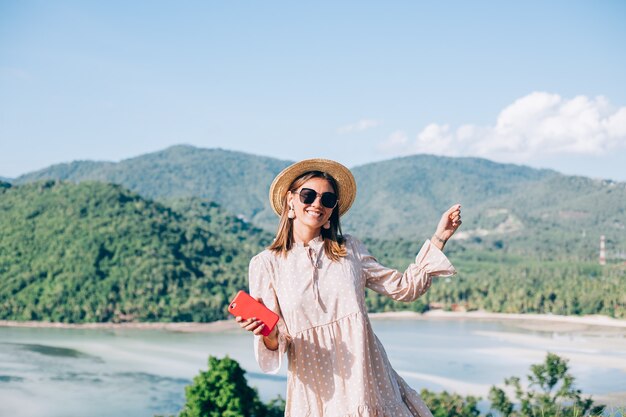 Mujer joven en vestido lindo de verano, sombrero de paja y gafas de sol bailando con smartphone en mano