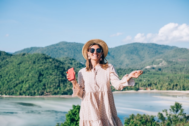 Mujer joven en vestido lindo de verano, sombrero de paja y gafas de sol bailando con smartphone en mano