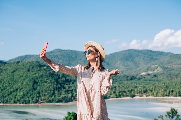 Mujer joven en vestido lindo de verano, sombrero de paja y gafas de sol haciendo videollamada con su teléfono inteligente