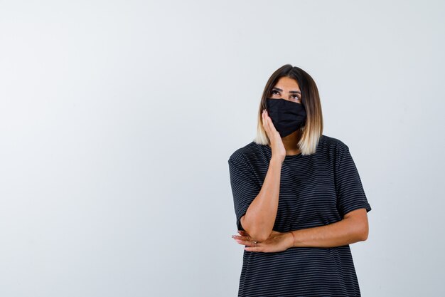 Mujer joven en vestido negro, máscara negra apoyada en la mejilla de la palma, pensando en algo y mirando pensativo, vista frontal.