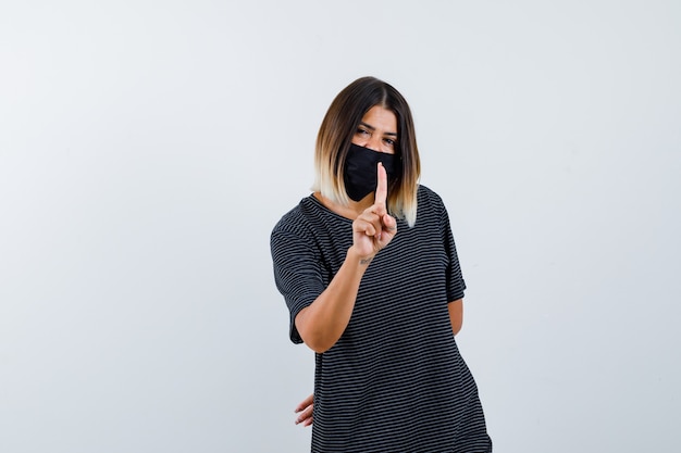 Foto gratuita mujer joven en vestido negro, máscara negra que muestra un gesto de un minuto, sosteniendo la mano detrás de la cintura y mirando feliz, vista frontal.
