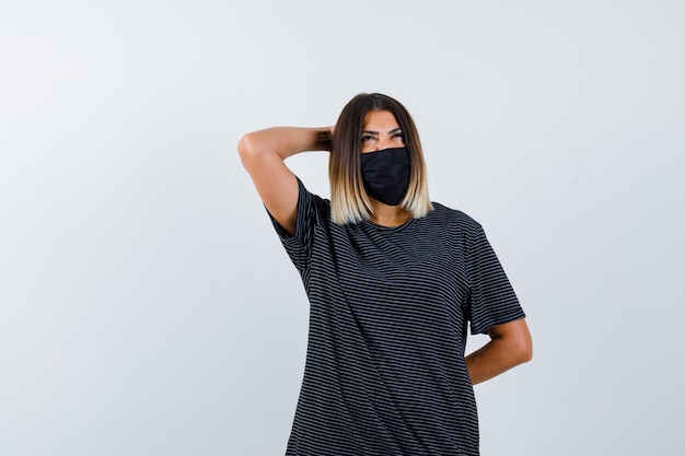 Mujer joven en vestido negro, máscara negra sosteniendo una mano detrás de la cabeza, otra mano detrás de la cintura, mirando hacia arriba y mirando pensativo, vista frontal.