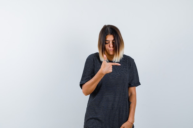 Foto gratuita mujer joven en vestido de polo apuntando hacia el lado derecho y mirando vacilante, vista frontal.