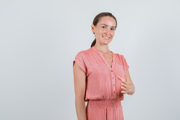 Mujer joven en vestido de rayas estirando la mano para agitar y mirar alegre, vista frontal.