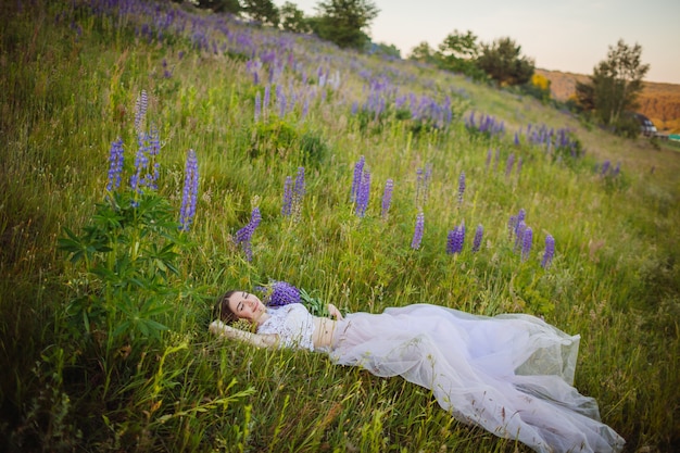 La mujer joven en vestido rico miente con el ramo de flores violetas en campo verde