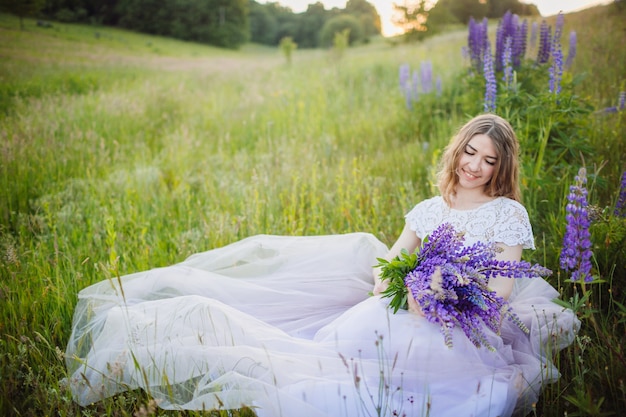 La mujer joven en vestido rico se sienta con el ramo de flores violetas en campo verde