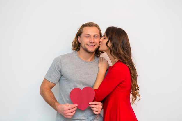 Mujer joven en vestido rojo que besa a su novio que sostiene el papel rojo de la forma del corazón contra el fondo blanco