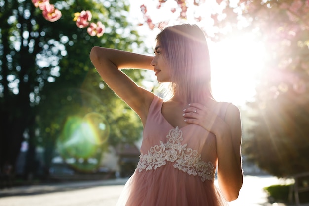 mujer joven en vestido rosa posa delante de un árbol de sakura lleno de flores rosadas e iluminado