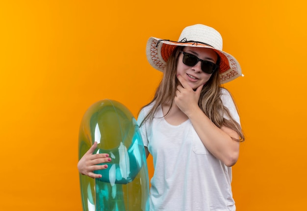 Foto gratuita mujer joven viajera en camiseta blanca con sombrero de verano con anillo inflable mirando con expresión pensativa en la cara de pie sobre la pared naranja