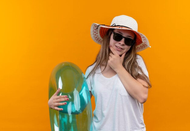 Mujer joven viajera en camiseta blanca con sombrero de verano con anillo inflable mirando con expresión pensativa en la cara de pie sobre la pared naranja