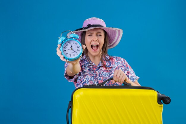 Mujer joven viajera con sombrero de verano de pie con maleta amarilla que muestra el despertador molesto y frustrado gritando con ira sobre fondo azul