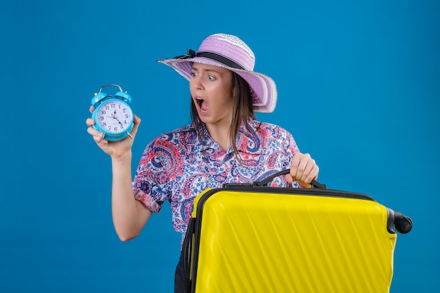 Foto gratuita mujer joven viajera con sombrero de verano de pie con maleta amarilla sosteniendo reloj despertador mirándolo sorprendido con vergüenza por error expresión de miedo sobre fondo azul