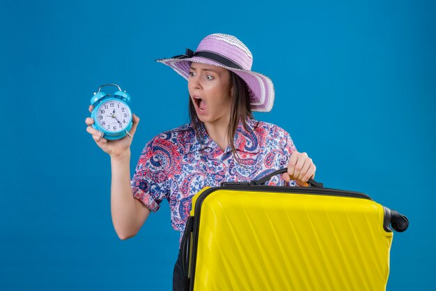 Mujer joven viajera con sombrero de verano de pie con maleta amarilla sosteniendo reloj despertador mirándolo sorprendido con vergüenza por error expresión de miedo sobre fondo azul